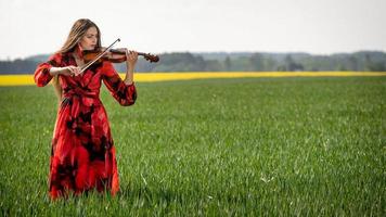 Young woman in red dress playing violin in green meadow - image photo