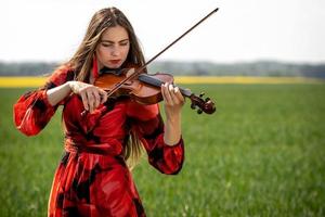 Young woman in red dress playing violin in green meadow - image photo