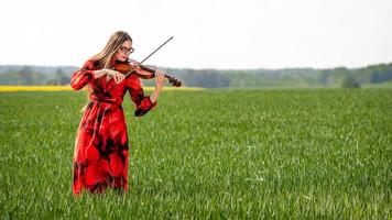 Young woman in red dress playing violin in green meadow - image photo