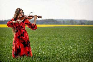 Young woman in red dress playing violin in green meadow - image photo