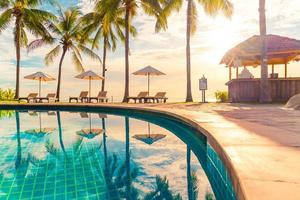 Beautiful luxury umbrellas and chairs around an outdoor swimming pool in hotel and resort with coconut palm trees on sunset or sunrise sky - holiday and vacation concept