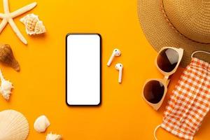 Fondo de verano con teléfono de pantalla en blanco y accesorios de playa, máscara para evitar el covid-19 en la vista superior de fondo naranja vibrante con espacio de copia. foto