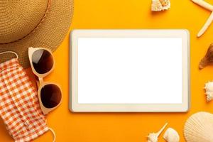 Summer background with blank screen tablet and beach accessories, mask to prevent covid-19 on vibrant orange background top view with copy space. photo