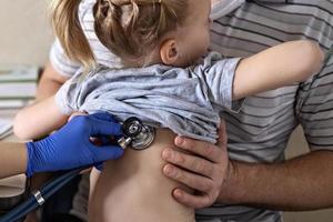 niña en los brazos de su padre en el consultorio del médico en la clínica. el médico examina al niño, escucha los pulmones con un fonendoscopio. tratamiento y prevención de infecciones respiratorias. foto