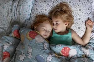 Two little sibling girls sisters sleeping in an embrace in bed under one blanket photo