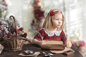 A little girl in a red dress is sitting at the table and writing a letter to Santa. Christmas card photo