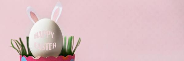 huevo de gallina blanco con orejas de conejo en una bandeja de papel rosa ecológico, caja. feliz concepto de vacaciones de pascua. texto en inglés feliz pascua .banner foto