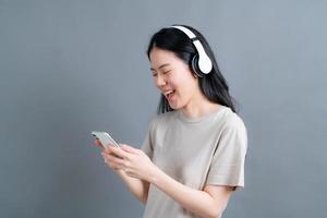 Asian woman wear wireless headphone hold smartphone looking at phone screen using mobile player app listening online music, learning foreign language, watching video relaxing on grey background photo