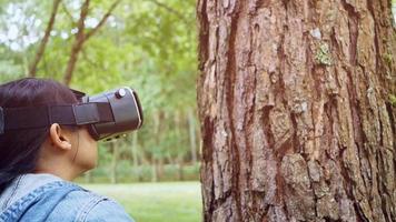 glad kvinna som bär VR-headsetglasögon av virtuell verklighet i skogen, berör det stora trädet på en solig sommardag i grön trädgård. modernt teknologikoncept. video