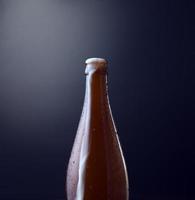 Front view of a bottle of cold beer with overflowing foam against blue grunge vintage background. Organic photography, vertical, square image style. photo