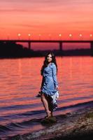 Beautiful young girl with long dark wavy hair standing at the bank of the river photo