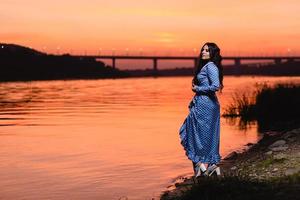 Hermosa joven con largo cabello ondulado oscuro de pie en la orilla del río foto