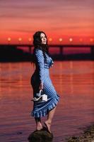 Beautiful young girl with long dark wavy hair standing at the bank of the river photo