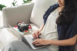 Fotógrafo de mujer asiática sentado en el sofá usando la computadora portátil para trabajar con cámara de película slr en casa, concepto creativo, empleado independiente. foto