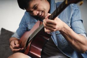 Disfrute de un apuesto hombre asiático practicando o tocando la guitarra en el sofá de la sala de estar