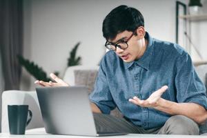 Joven asiático que usa una computadora portátil para una llamada de videoconferencia en línea, explique seriamente hablar en el sofá en la sala de estar por la noche. foto