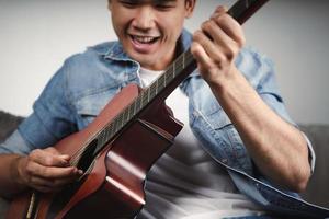 Disfrute de un apuesto hombre asiático practicando o tocando la guitarra en el sofá de la sala de estar