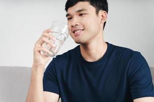 Apuesto hombre asiático bebiendo un vaso de agua en el sofá de la sala de estar