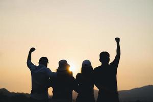 grupo de personas con los brazos levantados mirando al amanecer en el fondo de la montaña. conceptos de felicidad, éxito, amistad y comunidad. foto