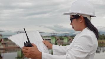 Female engineer in a white helmet reading data in a blueprint and using radio communication at the dam construction site to generate electricity. Clean energy and Technology concepts. video