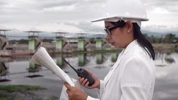 ingeniera con un casco blanco leyendo datos en un plano y usando comunicación por radio en el sitio de construcción de la presa para generar electricidad. conceptos de energía y tecnología limpias. video