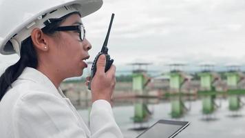 vrouwelijke ingenieur in een witte helm die gegevens in een blauwdruk leest en radiocommunicatie gebruikt op de bouwplaats van de dam om elektriciteit op te wekken. schone energie en technologieconcepten. video