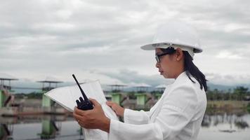 femme ingénieur dans un casque blanc lisant des données dans un plan et utilisant la communication radio sur le chantier de construction du barrage pour produire de l'électricité. concepts d'énergie propre et de technologie. video