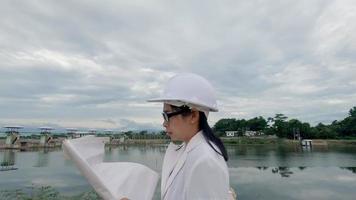 Female engineer in a white helmet reading data in a blueprint and using radio communication at the dam construction site to generate electricity. Clean energy and Technology concepts. video