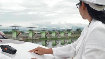 ingeniera con un casco blanco leyendo datos en un plano y usando comunicación por radio en el sitio de construcción de la presa para generar electricidad. conceptos de energía y tecnología limpias. video