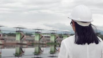 ingeniera con un casco blanco trabajando con una tableta digital y mirando hacia otro lado en el sitio de construcción de la presa para generar electricidad. video