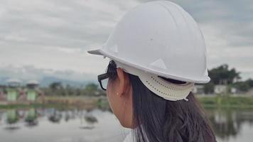 ingeniera con un casco blanco trabajando con una tableta digital y mirando hacia otro lado en el sitio de construcción de la presa para generar electricidad.