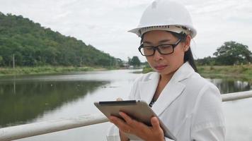 engenheira em um capacete branco, trabalhando com um tablet digital e olhando para o local de construção da barragem para gerar eletricidade.