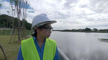 ingeniera con un casco blanco y uniforme amarillo sostiene un plano y mirando hacia otro lado en el sitio de construcción de la presa para generar electricidad.