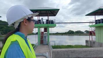 ingegnere donna con un casco bianco e un'uniforme gialla tiene un progetto e guarda il cantiere della diga per generare elettricità. video