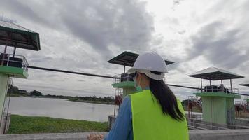 vrouwelijke ingenieur in een witte helm en geel uniform houdt een blauwdruk vast en kijkt weg naar de bouwplaats van de dam om elektriciteit op te wekken. video