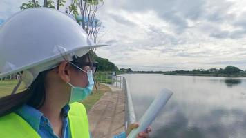 vrouwelijke ingenieur in een witte helm en geel uniform houdt een blauwdruk vast en kijkt weg naar de bouwplaats van de dam om elektriciteit op te wekken. video