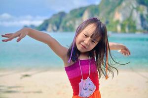 Cute portraits of black-haired smiling Asian girl braided with multicolored thread in casual rainbow dress and face mask, beautiful beach with mountain and sea in summer holiday vacation. photo