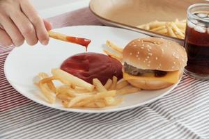Close-up shot, selective focus. Man's hand was eating French fried, dipping ketchup on a white plate on red tablecloth with hamburgers and cola. Eating junk food or fast food for lunch is unhealthy. photo