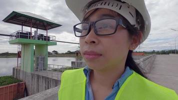 vrouwelijke ingenieur in een witte helm en geel uniform houdt een blauwdruk vast en kijkt weg naar de bouwplaats van de dam om elektriciteit op te wekken. video