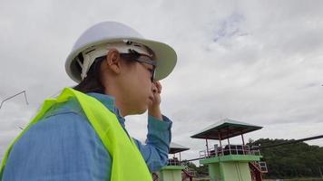 Female engineer in a white helmet and yellow uniform holds a blueprint and looking away at the dam construction site to generate electricity. video