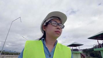 ingegnere donna con un casco bianco e un'uniforme gialla tiene un progetto e guarda il cantiere della diga per generare elettricità. video