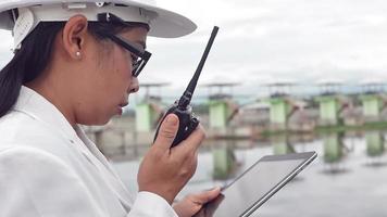 ingeniera con un casco blanco trabajando con una tableta digital y mirando hacia otro lado en el sitio de construcción de la presa para generar electricidad. video