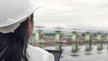 Female engineer in a white helmet working with a digital tablet and looking away at the dam construction site to generate electricity. video