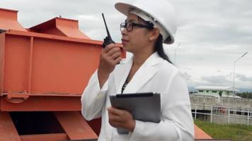 ingeniera con un casco blanco sostiene una tableta digital y utiliza la comunicación por radio en el sitio de construcción de la presa para generar electricidad. conceptos de energía y tecnología limpias. video