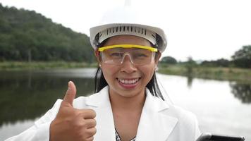 Female engineer in a helmet looks and smiles at the camera while holding a digital tablet with her thumb up at a dam construction site to generate electricity. video