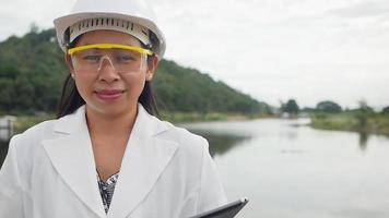 Female engineer in a helmet looks and smiles at the camera while holding a digital tablet with her thumb up at a dam construction site to generate electricity. video