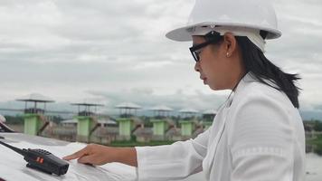 ingeniera con un casco blanco leyendo datos en un plano y usando comunicación por radio en el sitio de construcción de la presa para generar electricidad. conceptos de energía y tecnología limpias. video