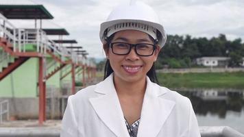 ingeniera con un casco blanco mira y sonríe a una cámara en el sitio de construcción de una presa para generar electricidad. video