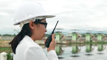 Female engineer in a white helmet using radio communication with worker at the dam construction site to generate electricity. video