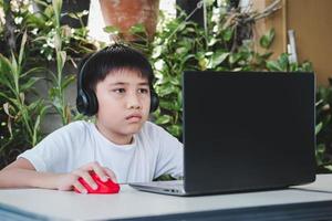 Asian boy wears wireless headphones and uses laptop computer to study online photo
